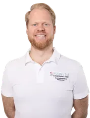 Smilende mann med rødt skjegg og blondt hår, iført en hvit poloskjorte med logo på skjorten.